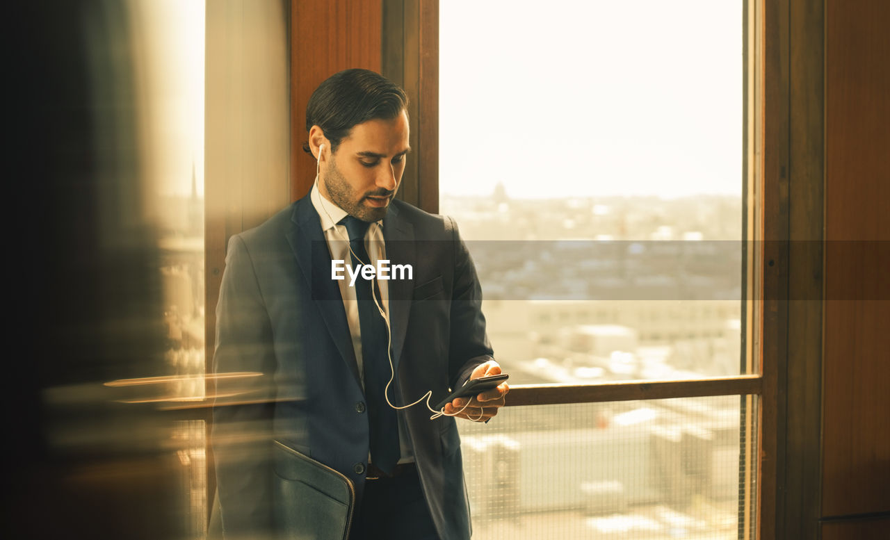 Mid adult legal professional using smart phone while standing against window at law office