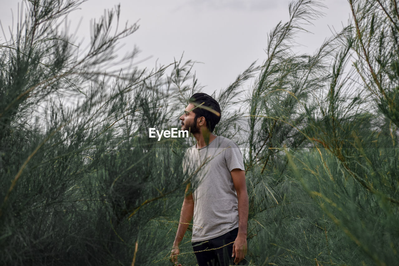 Young man looking away while standing on land against trees