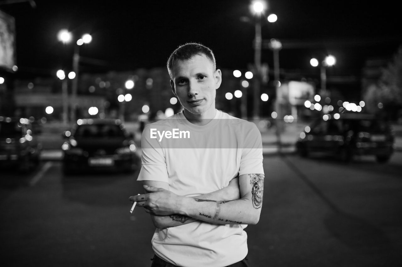 FULL LENGTH PORTRAIT OF YOUNG MAN ON ILLUMINATED STREET AT NIGHT