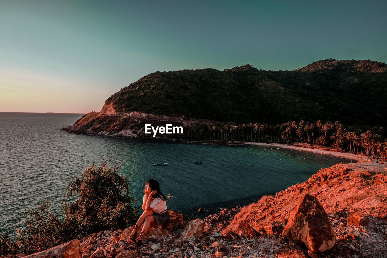 Full length of girl sitting on rock by sea against sky