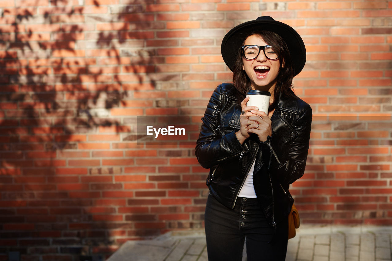brick, brick wall, wall, adult, one person, young adult, clothing, fashion, standing, emotion, happiness, smiling, women, jacket, enjoyment, wall - building feature, person, photo shoot, lifestyles, city, leather jacket, fun, red, arts culture and entertainment, singing, portrait, music, architecture, three quarter length, holding, cheerful, front view, leather, human mouth, leisure activity, copy space, city life, microphone, casual clothing, fashion accessory, hat, mouth open, input device, black