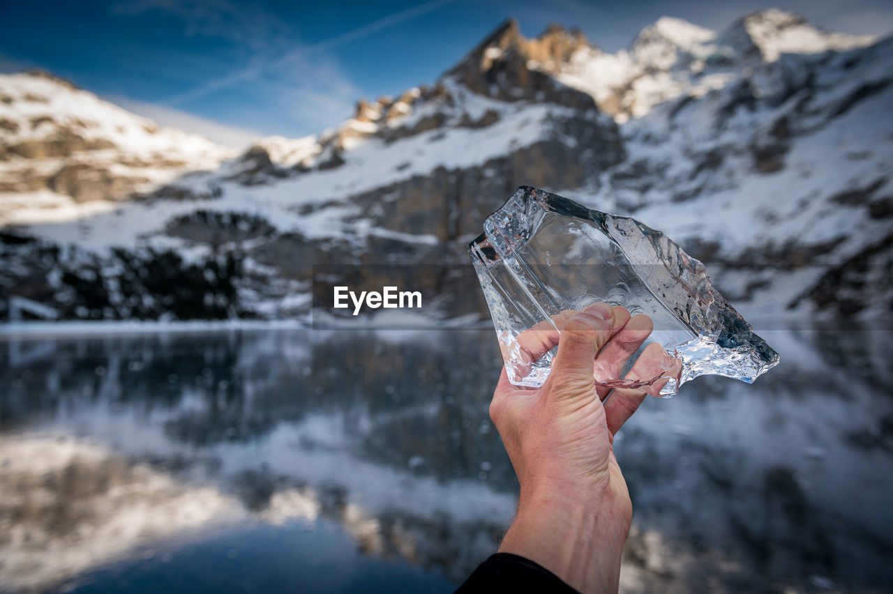 Cropped image of person holding ice by lake against mountains and sky