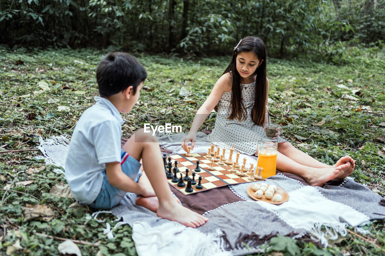 Barefoot girl with brother playing chess while sitting on textile with jug of drink and biscuits on lawn