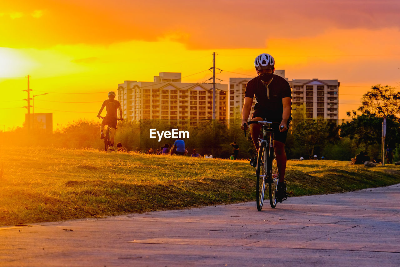 transportation, bicycle, sky, sunset, sports, cycling, architecture, city, evening, activity, nature, helmet, one person, headwear, men, adult, riding, road, motion, mode of transportation, built structure, lifestyles, full length, sports helmet, building exterior, orange color, vehicle, cloud, leisure activity, land vehicle, street, sports equipment, sunlight, outdoors, on the move, exercising