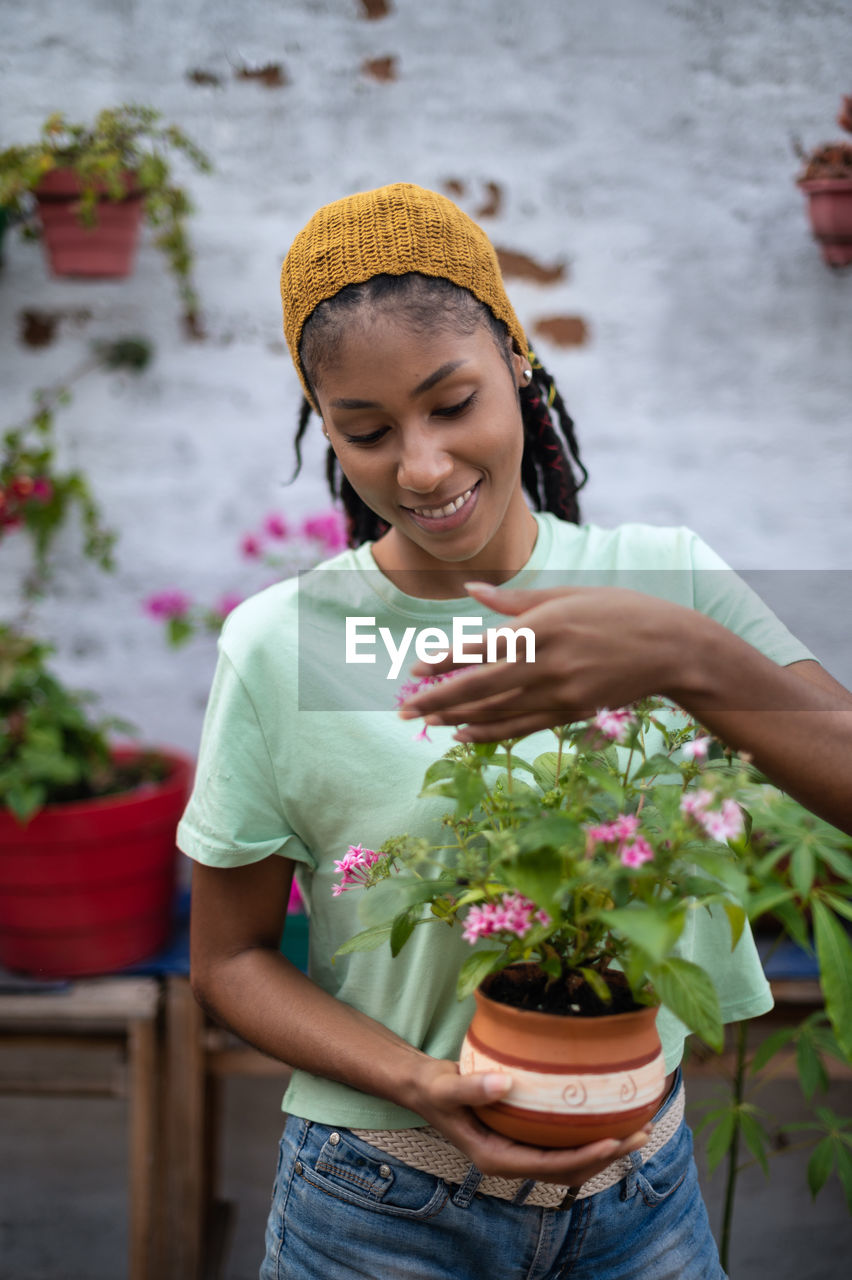 Delighted african american female gardener enjoying scent of pentas lanceolata flower while standing in greenhouse and looking at camera