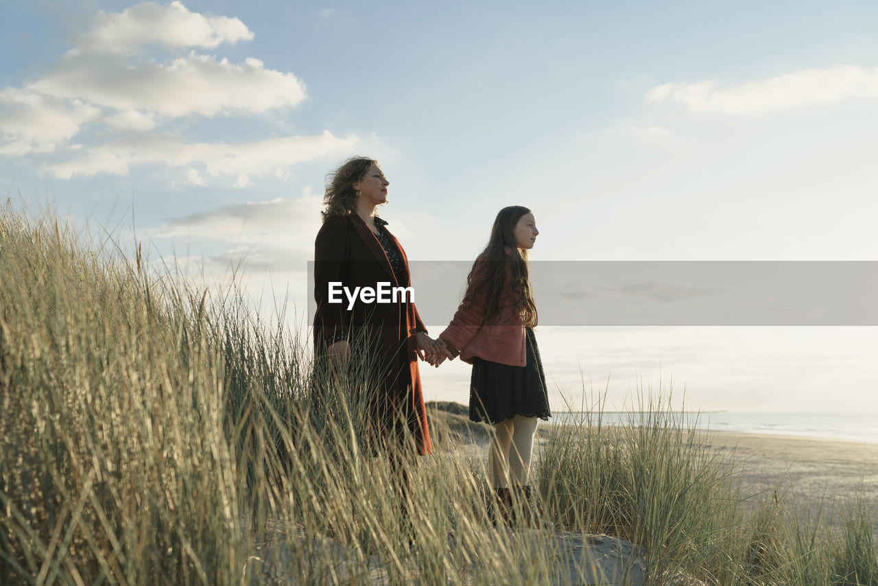 Woman with daughter standing on grass at beach against sky