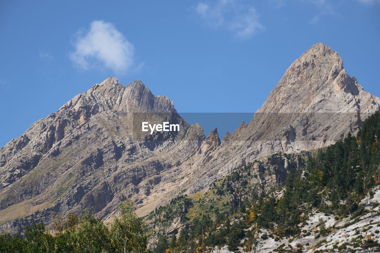 Grassy valley with bushes near rough rocky mountain range located against blue sky in nature on sunny day in valle de pineta in pyrenees, spain