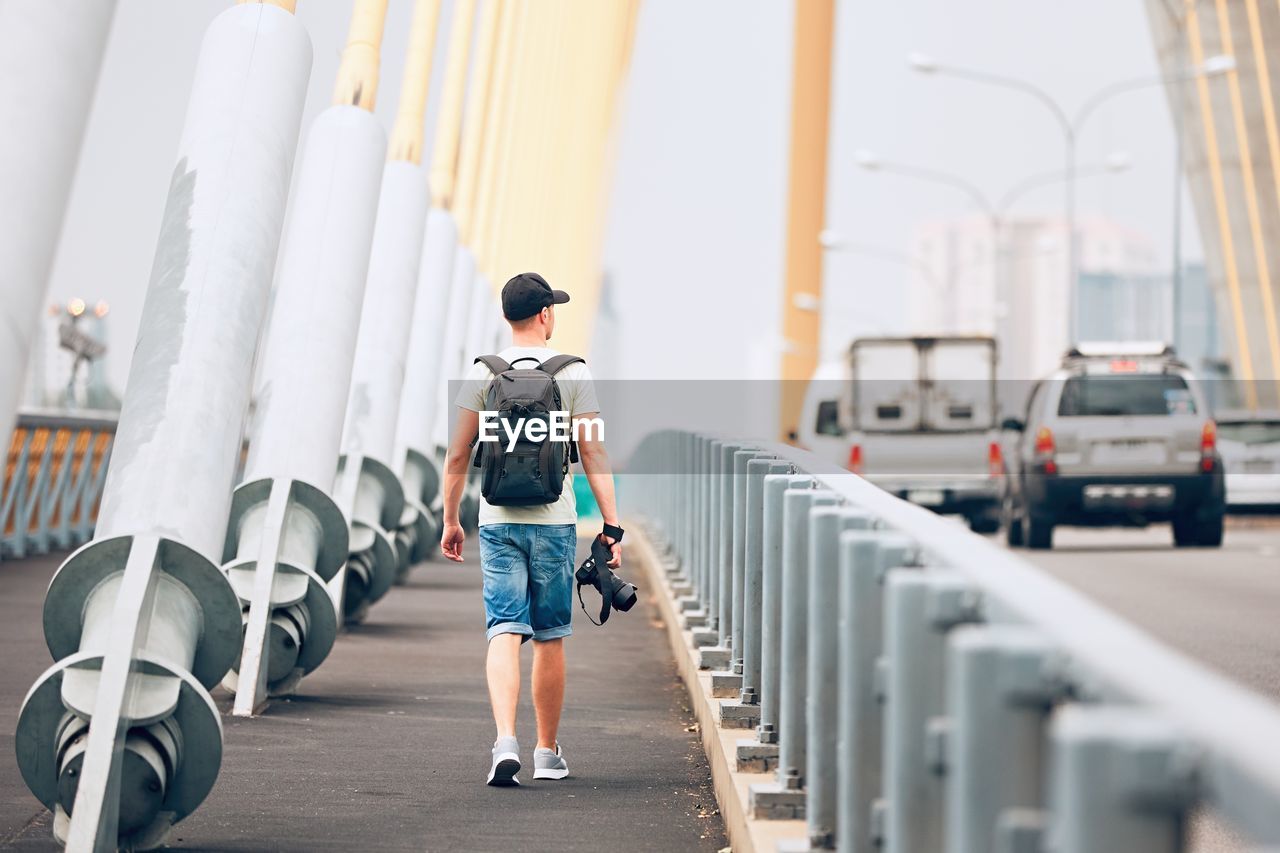 Rear view of young man holding camera while walking on bridge against sky