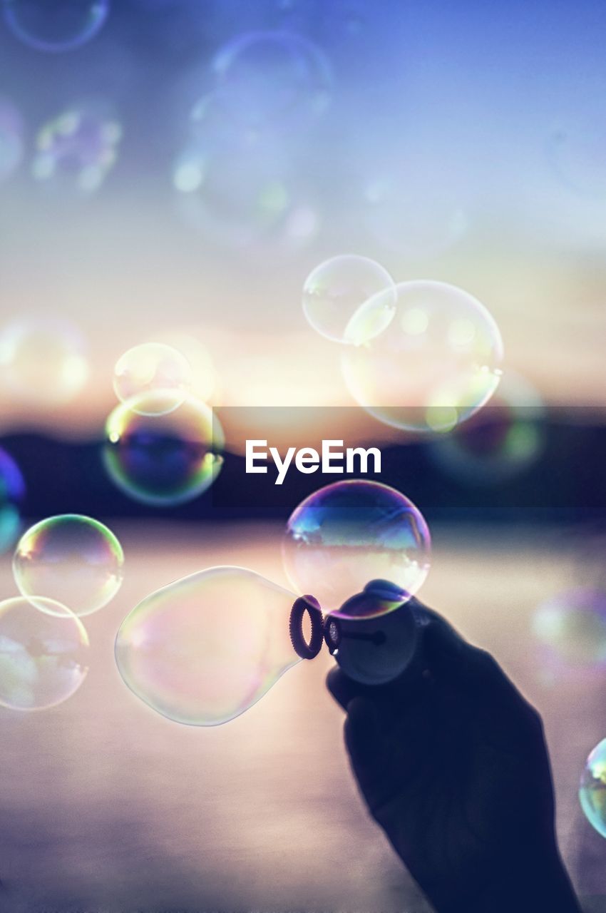 Cropped hand of man making bubbles by lake during sunset