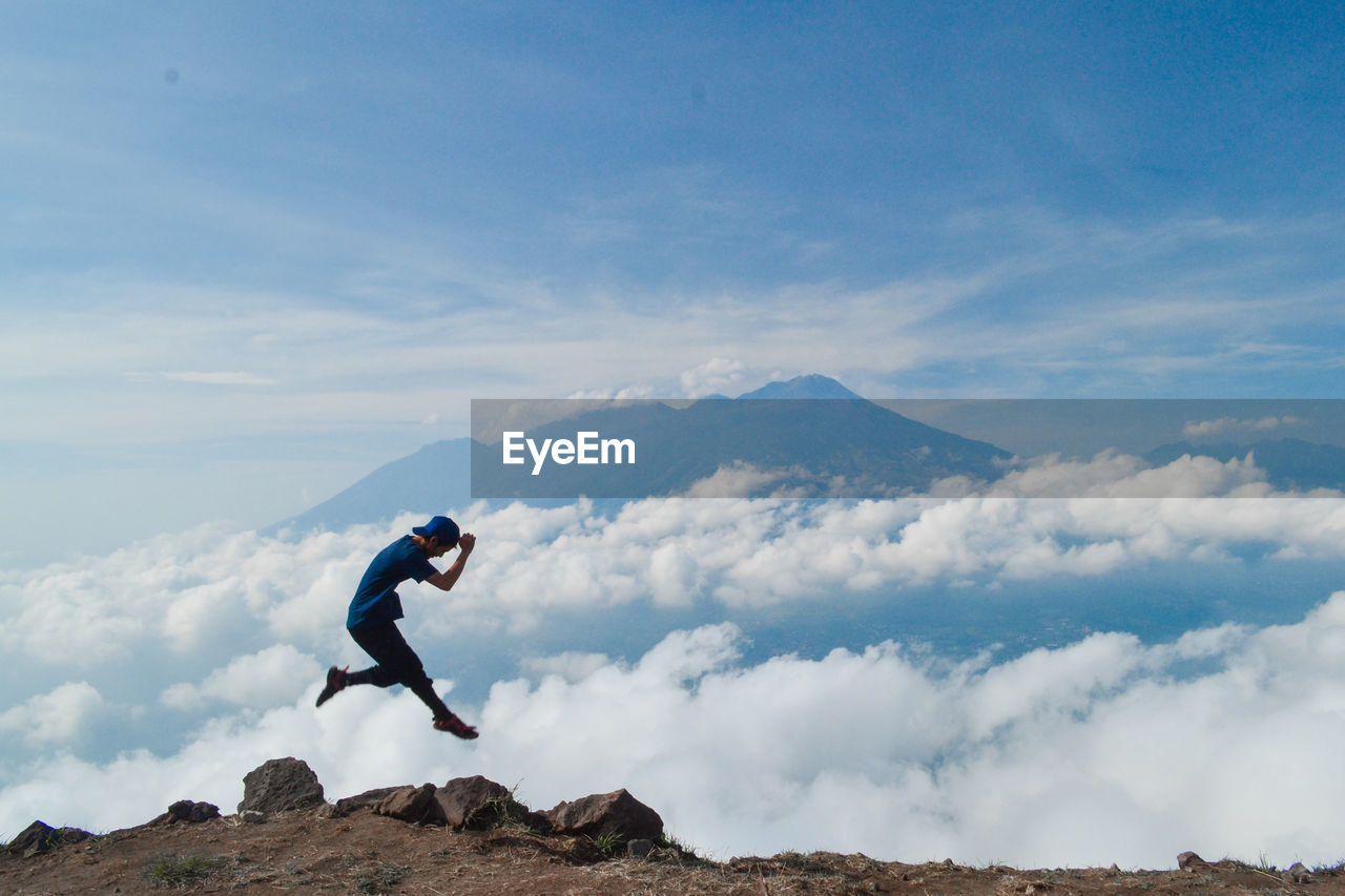 Side view of hiker jumping on mountain peak against sky