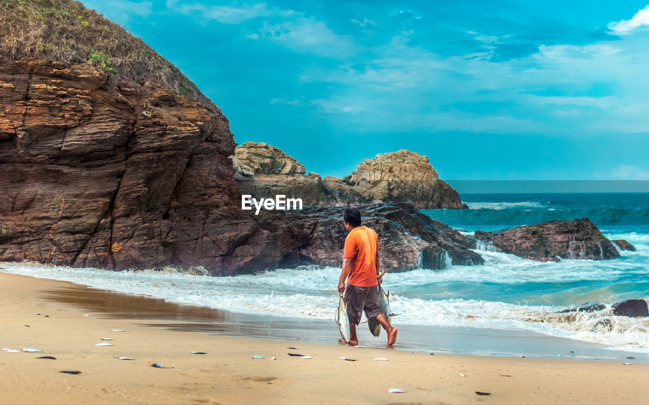 Rear view of man with fishes walking at beach against cloudy sky