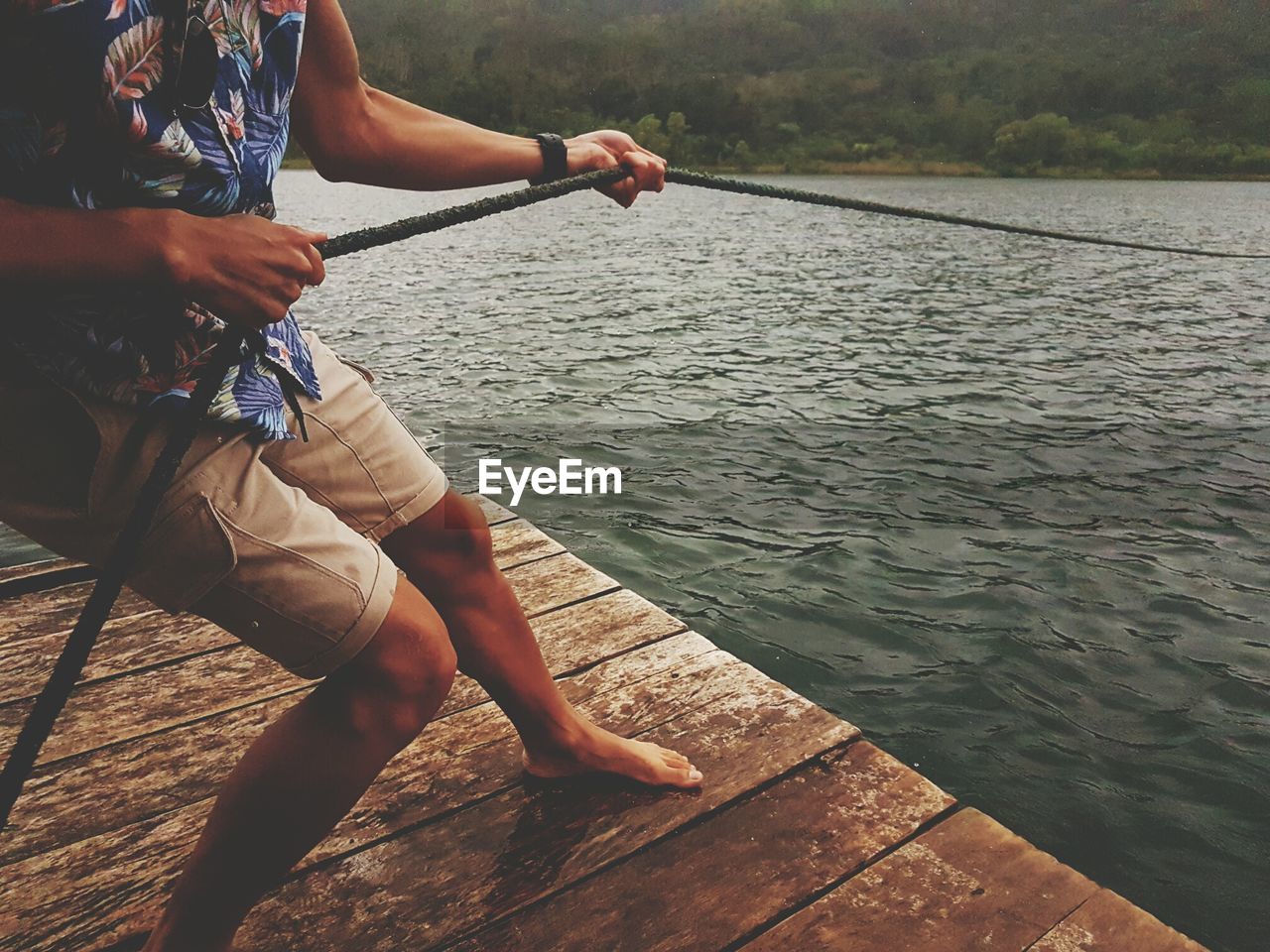 Low section of man pulling rope while standing on pier over lake