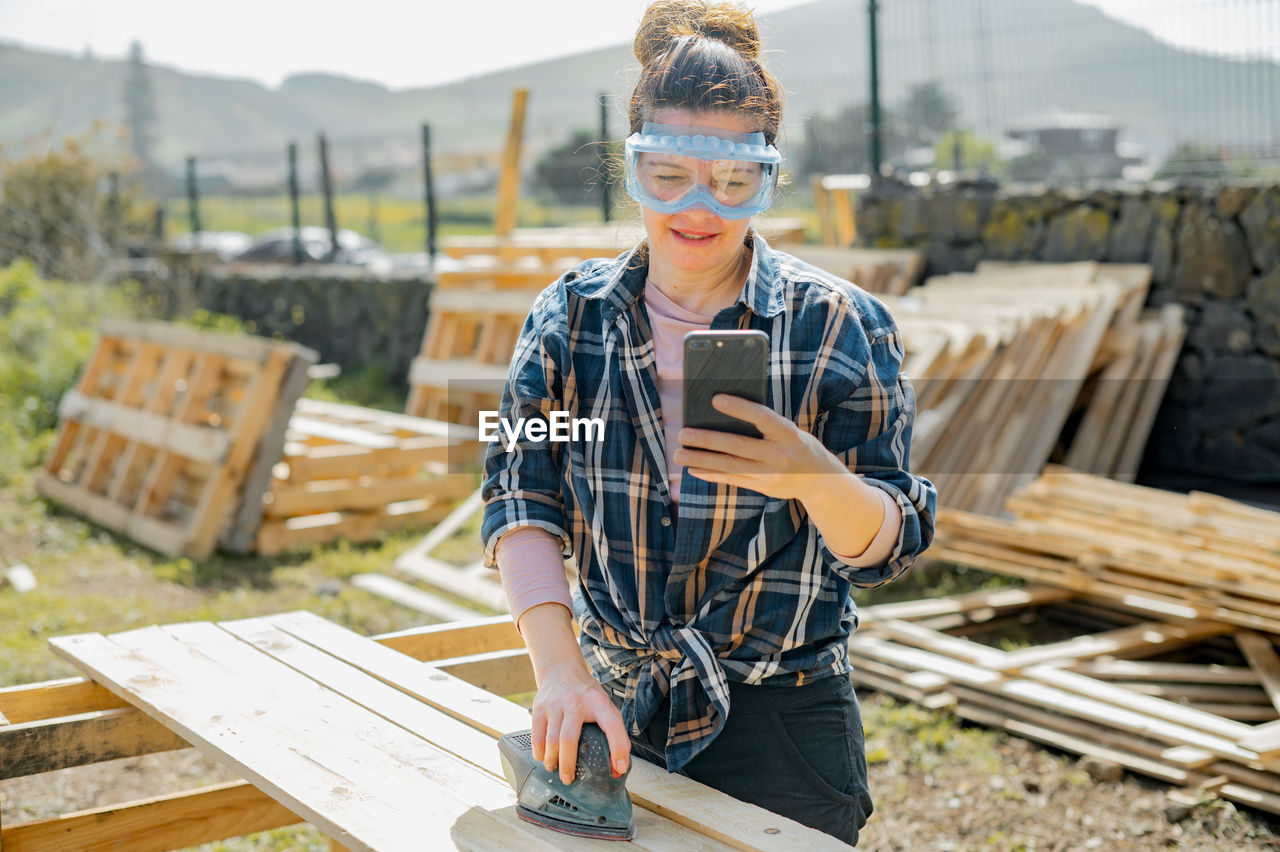 Woman in protective glasses text messaging on cellphone while sanding down wooden planks with orbit sander in countryside on summer day