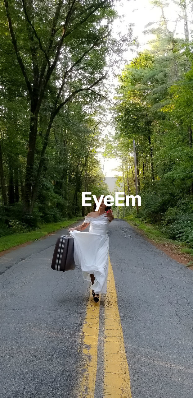 Woman holding suitcase while running on road amidst trees