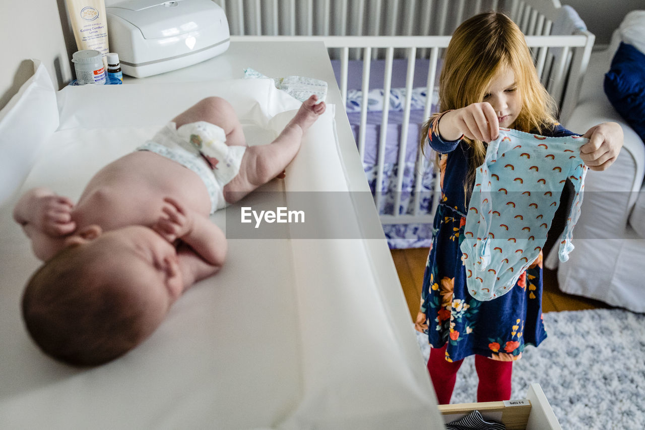 Girl choosing clothes for shirtless baby sister lying on table at home