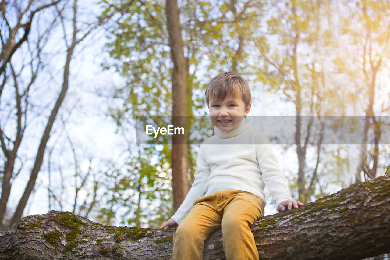 PORTRAIT OF HAPPY BOY SITTING ON TREE TRUNK