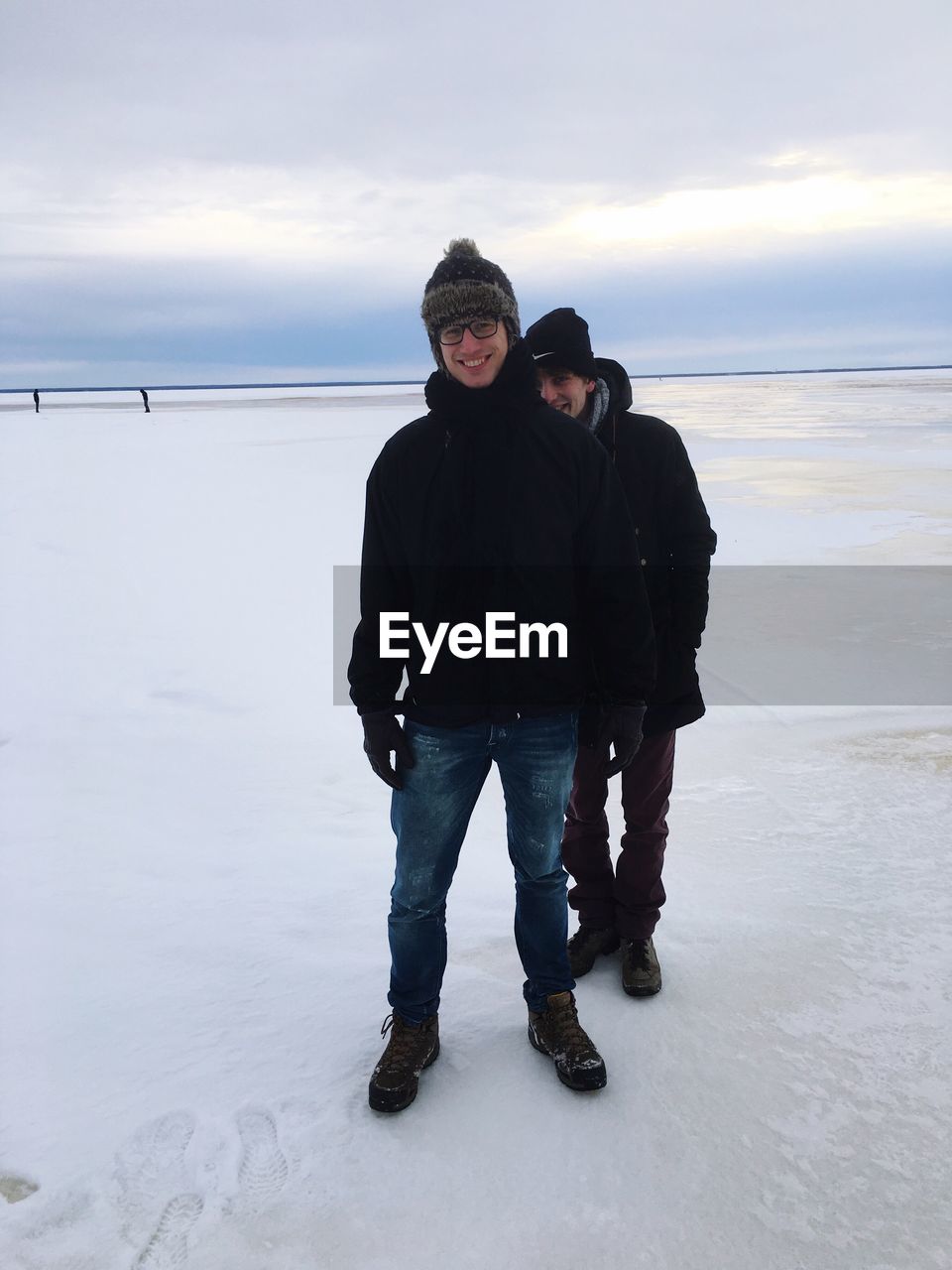 Smiling friends standing on snow covered field against cloudy sky