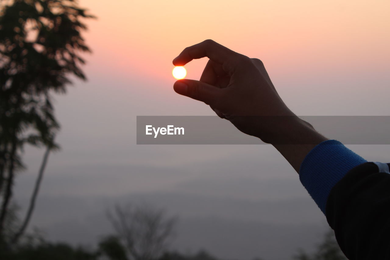 Optical illusion of cropped hand holding sun against sky during sunset