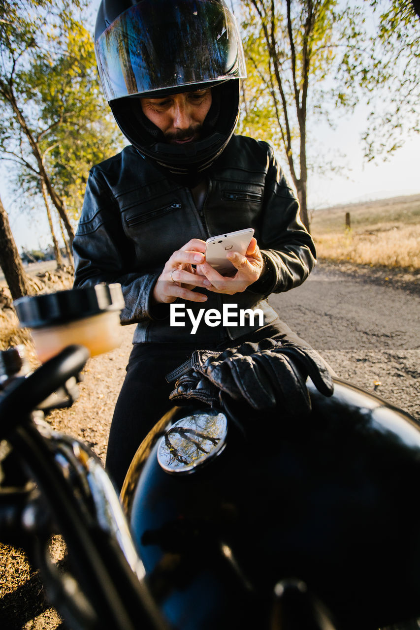 Concentrated male racer in leather jacket sitting on motorbike and browsing phone in autumn sunny day