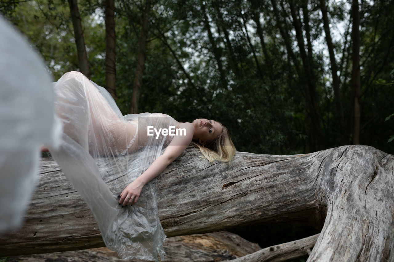 Portrait of naked woman wrapped in a polythene lying down on tree trunk in forest