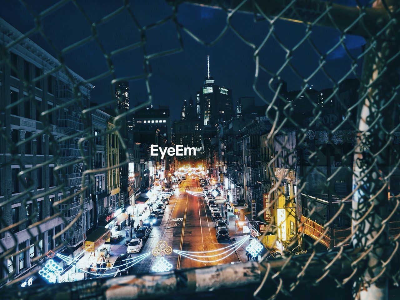 Vehicles parked on illuminated street amidst buildings seen through metallic fence at night