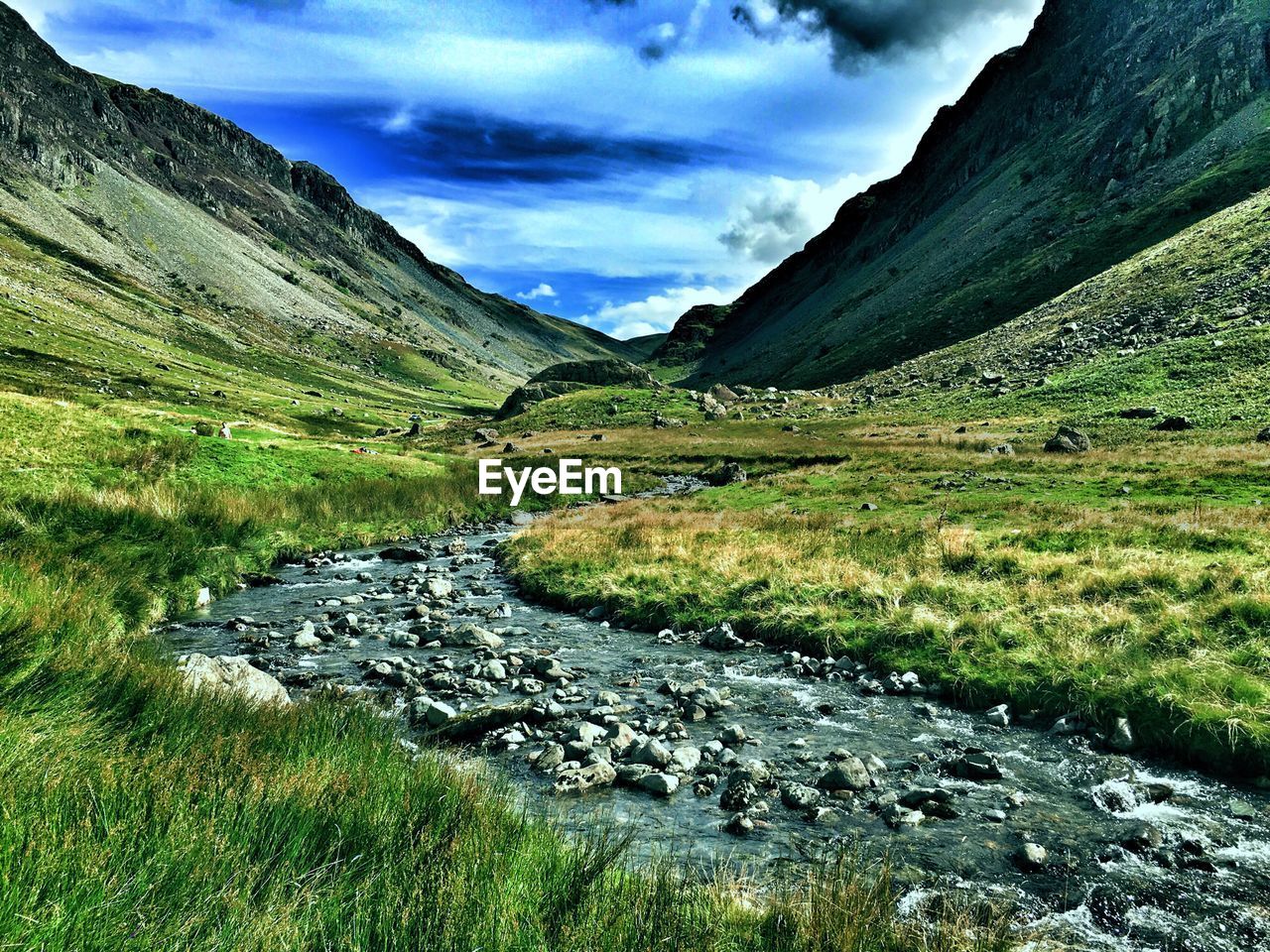 Scenic view of stream flowing on grassy field by mountains against sky