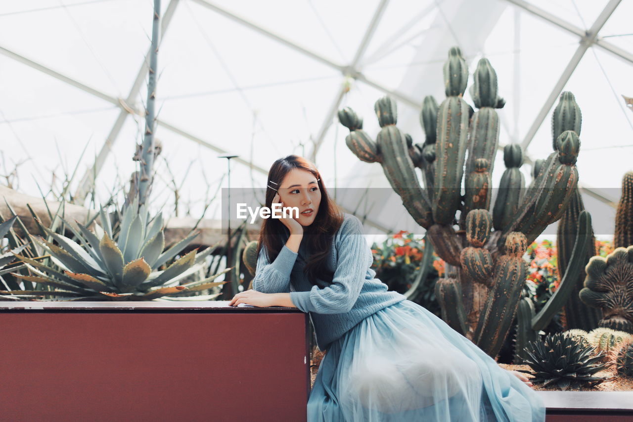 Thoughtful young woman sitting against plants in greenhouse