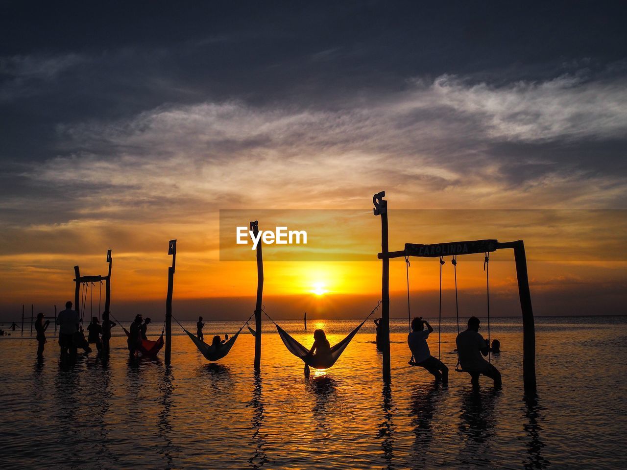 Silhouette people resting on swings and hammocks over sea against sky at sunset