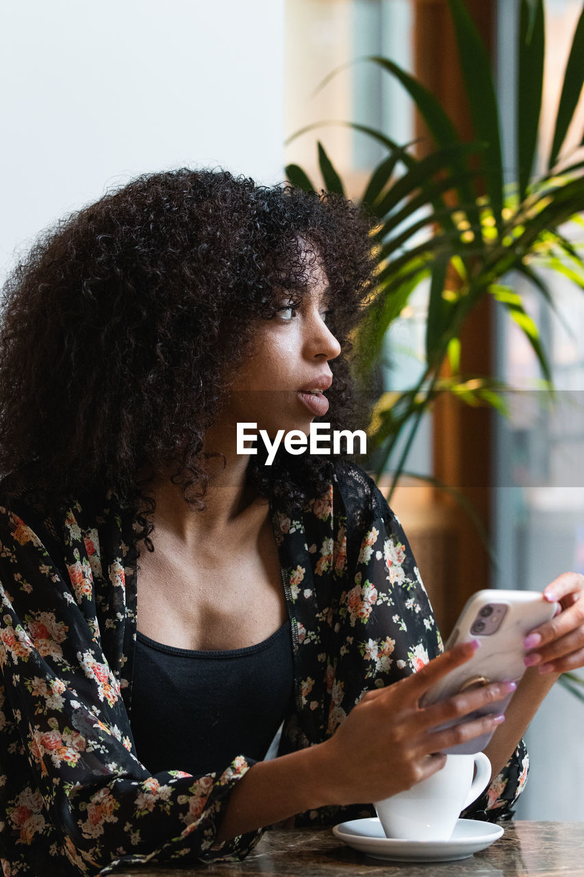 Young ethnic female browsing on cellphone while sitting looking away at table with a cup of coffee