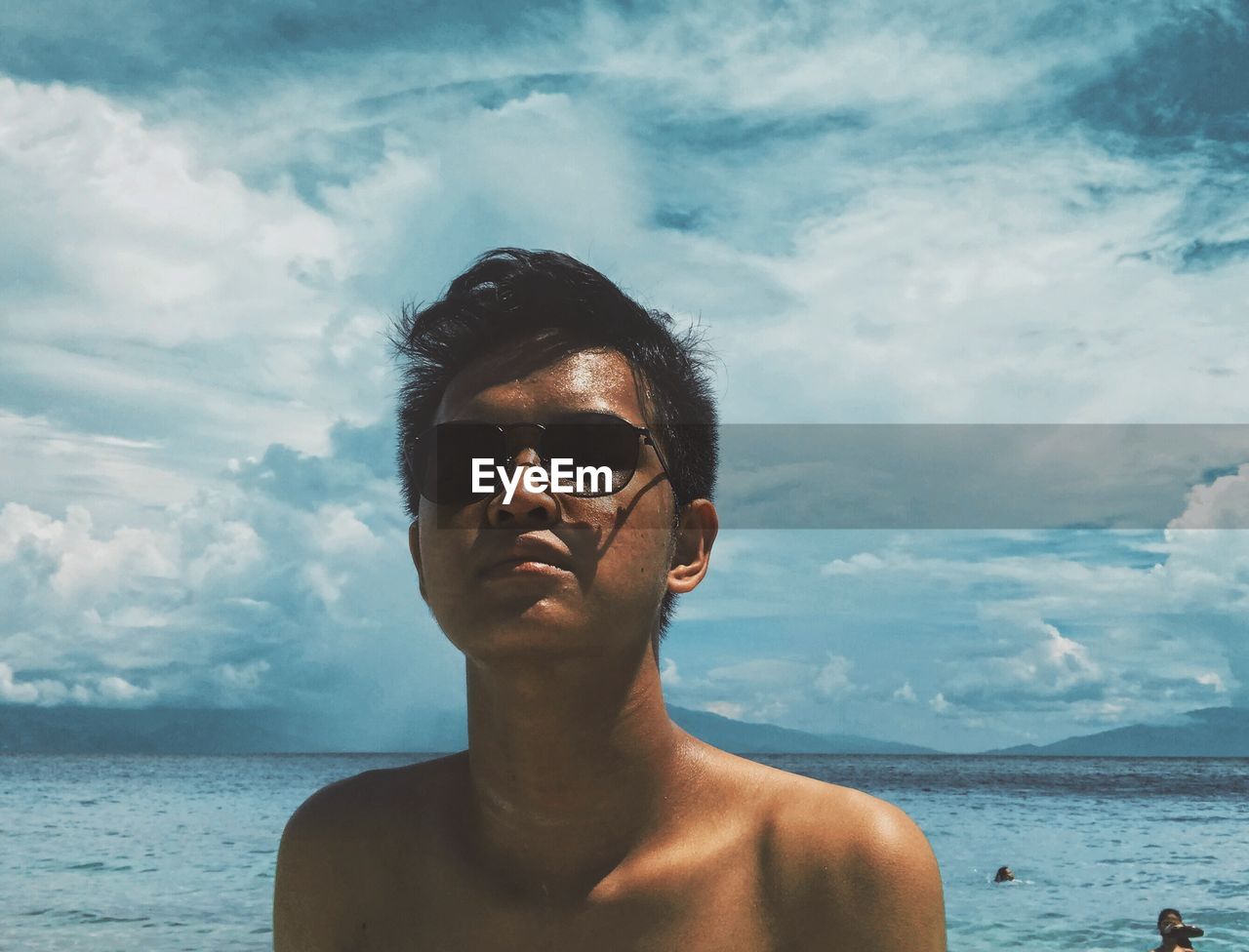 Shirtless young man wearing sunglasses at beach against cloudy sky
