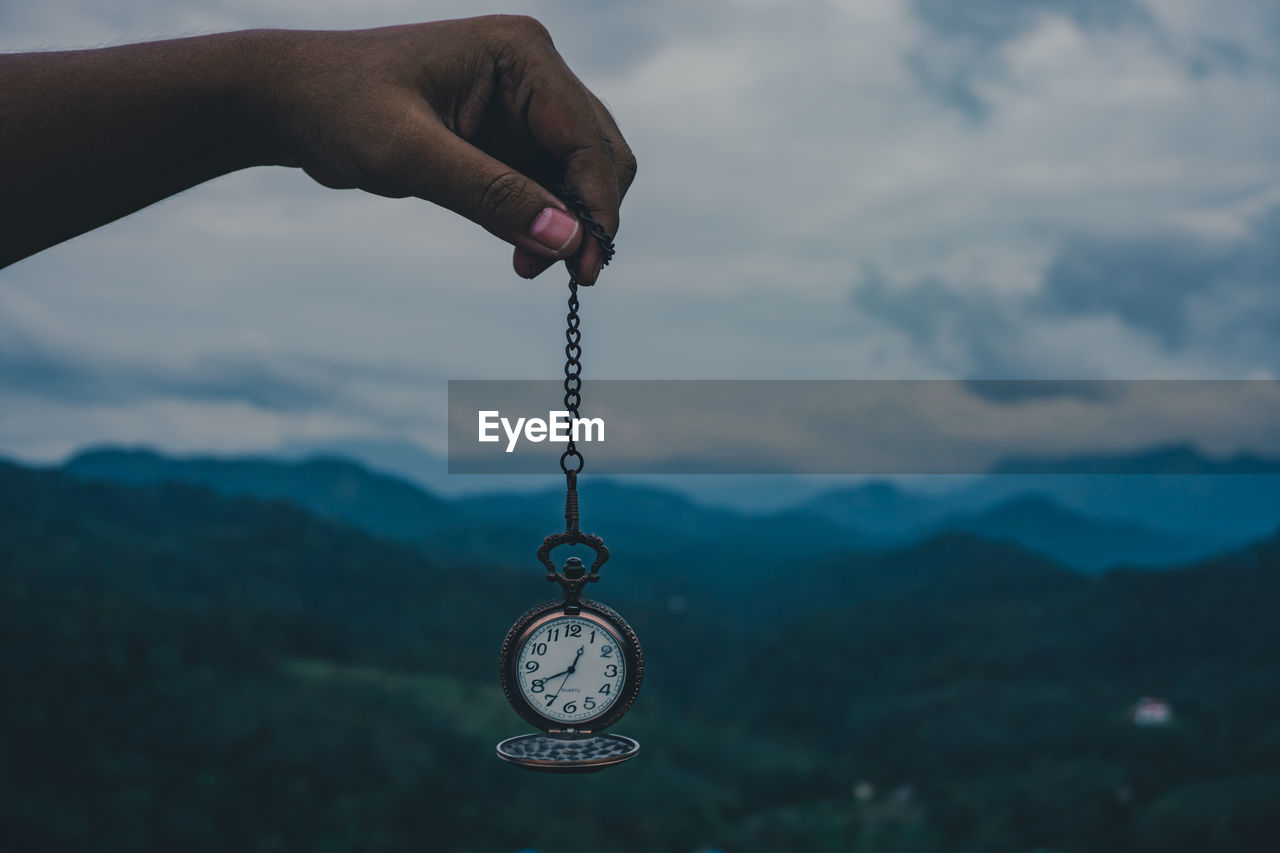 CLOSE-UP OF HAND HOLDING CLOCK AGAINST CLOUDS