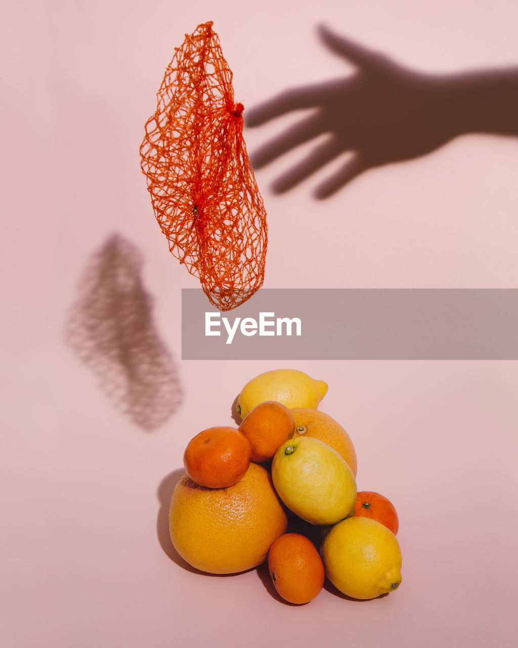 Still life with citrus fruits, orange net floating and the shadow of a hand on a pink background