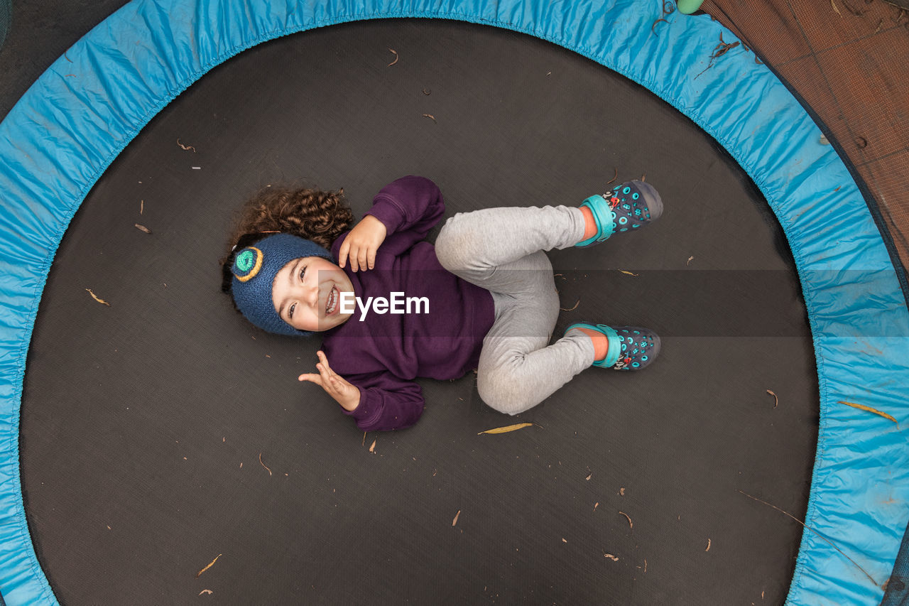 Top view full body of joyful ethnic child looking at camera while lying on trampoline and having fun at daytime