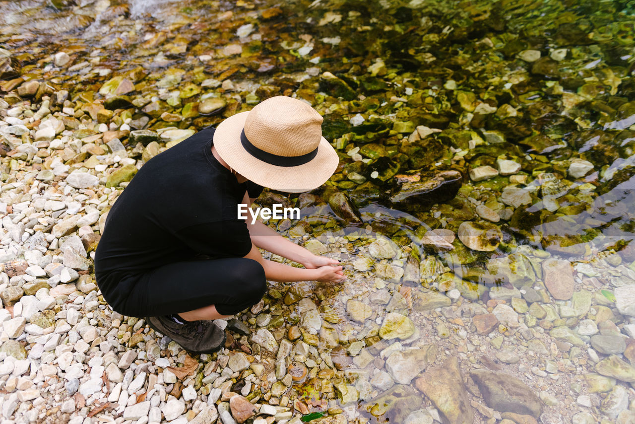 From above of female tourist drinking clear water from calm river in forest during summer adventure