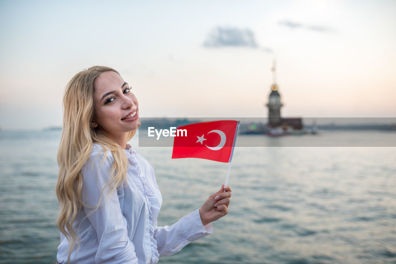 Portrait of young woman with blond hair holding turkish flag against sea