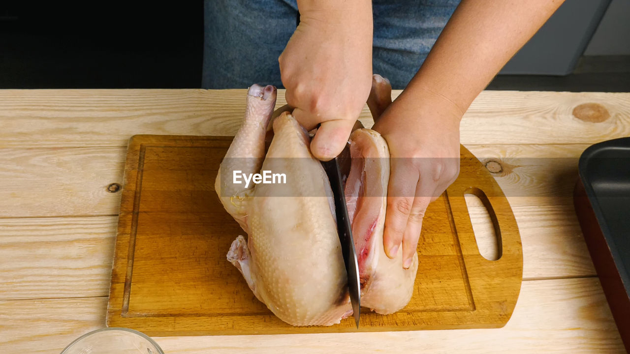 A woman is cutting a raw chicken carcass in half on a cutting board with a large knife.