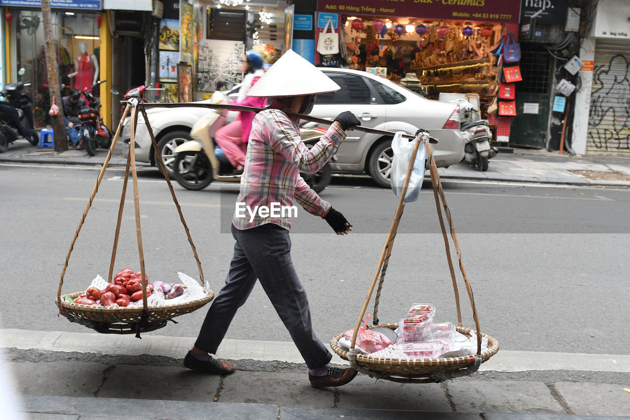 road, transportation, city, street, adult, mode of transportation, one person, full length, architecture, women, vehicle, day, casual clothing, city life, umbrella, infrastructure, holding, men, retail, building exterior, occupation, lifestyles, hat, food, market, built structure, small business, outdoors, standing, vendor, travel, clothing, business, fashion accessory, car, person, selling, city street