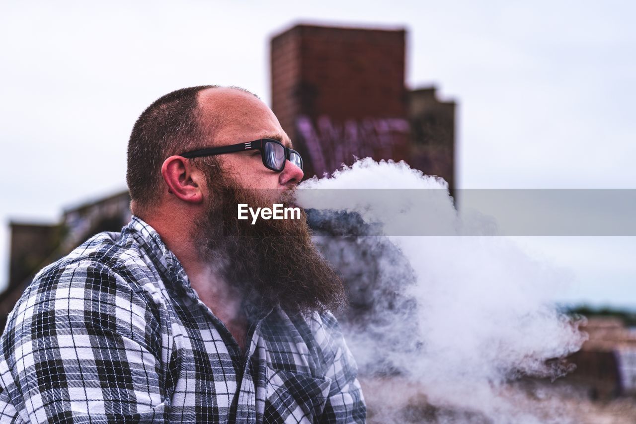 Man exhaling smoke while sitting against sky