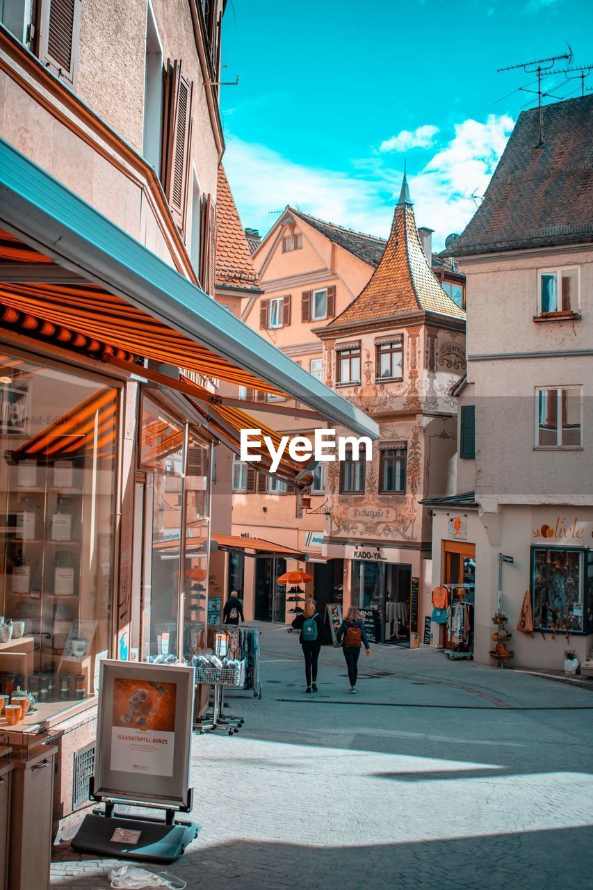 PEOPLE WALKING ON STREET AMIDST BUILDINGS IN CITY AGAINST SKY