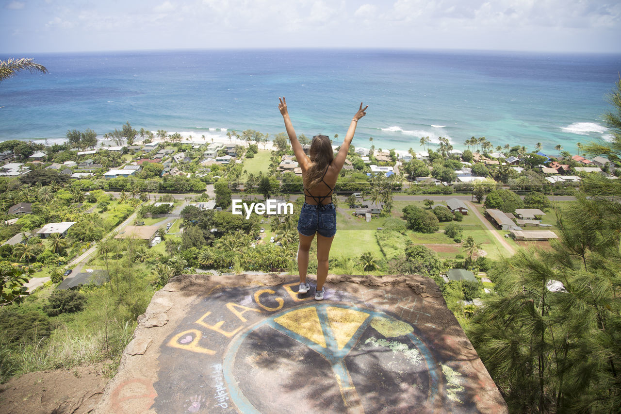 Full length of woman standing with hands up with view over sea and grafitti