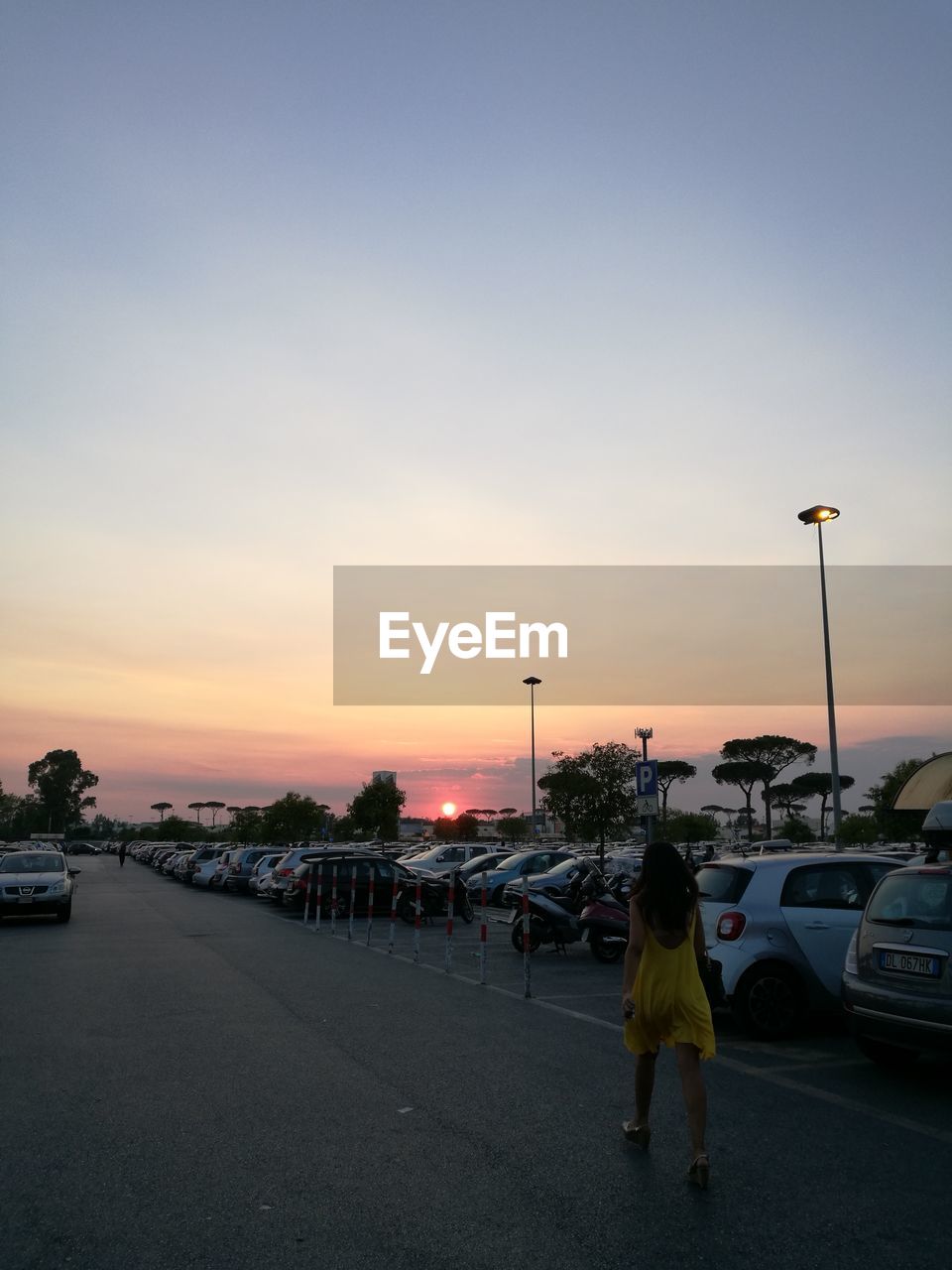 REAR VIEW OF WOMAN STANDING BY CAR AGAINST SKY DURING SUNSET