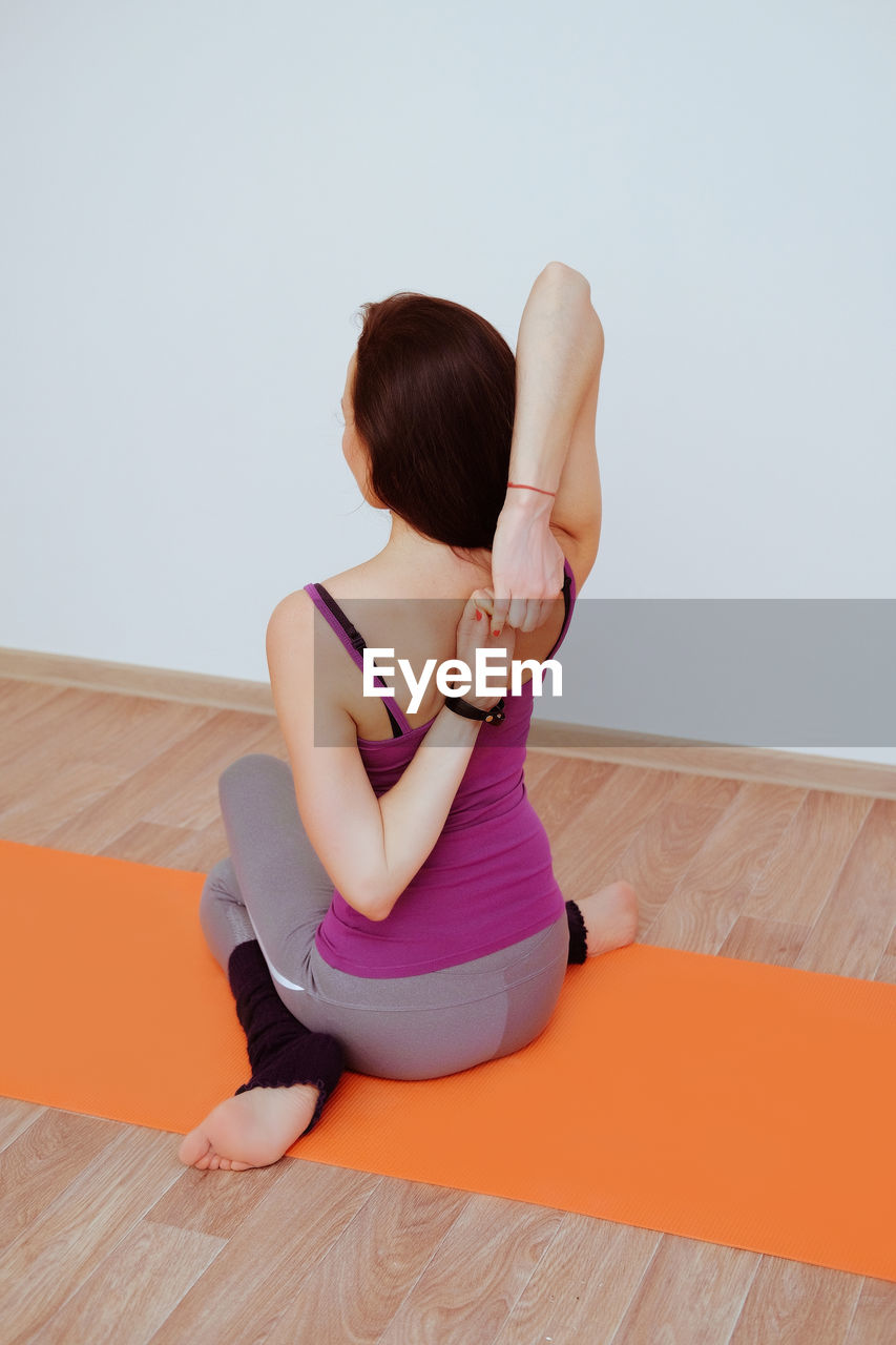 REAR VIEW OF WOMAN SITTING ON HARDWOOD FLOOR AGAINST WHITE WALL