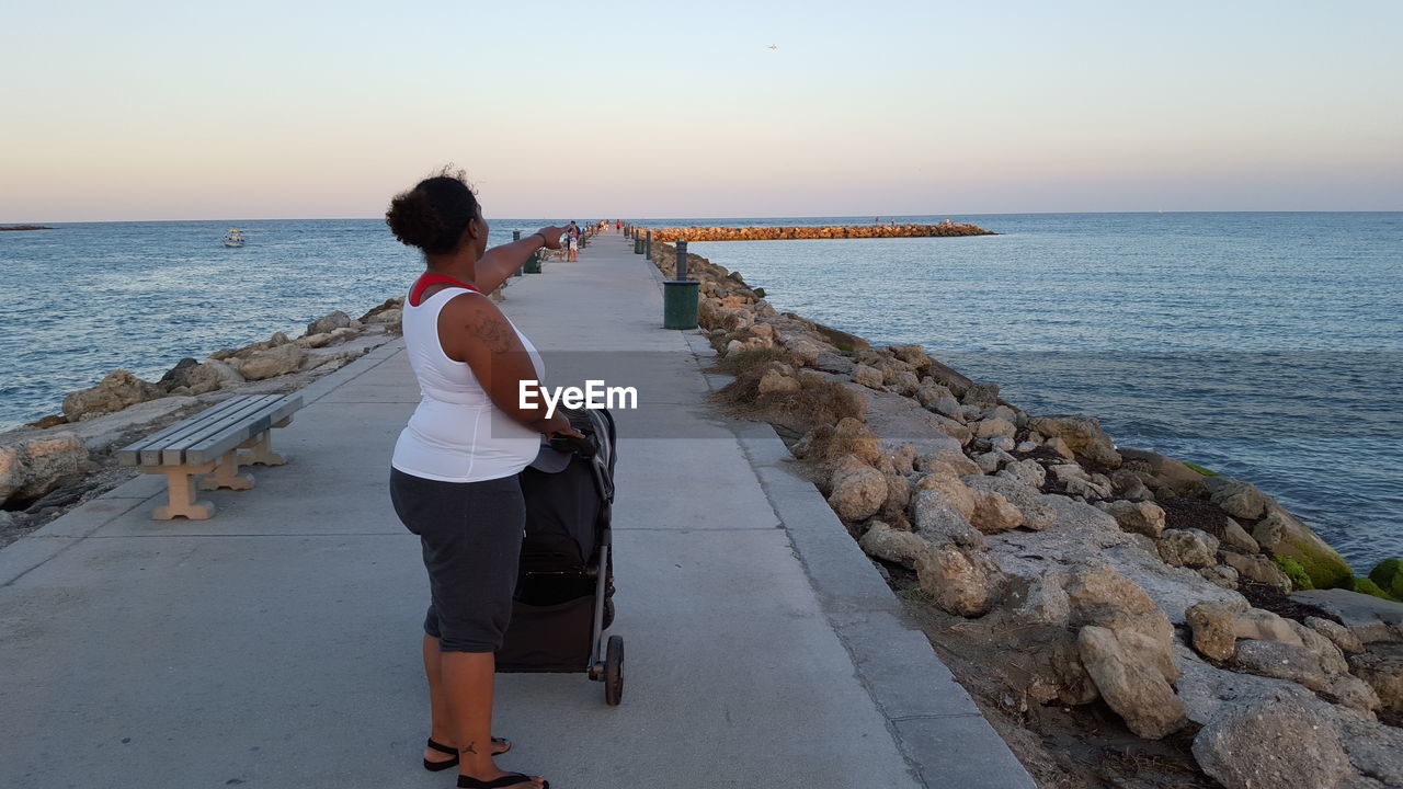 Side view of woman with baby carriage pointing away at pier over sea