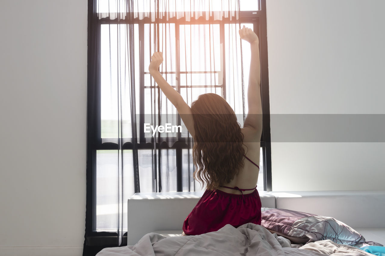 REAR VIEW OF WOMAN STANDING BY WINDOW