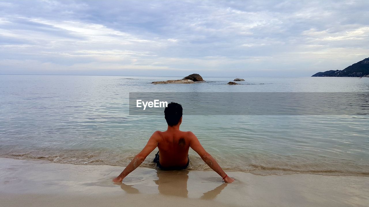 Rear view of shirtless man resting on shore at beach against cloudy sky