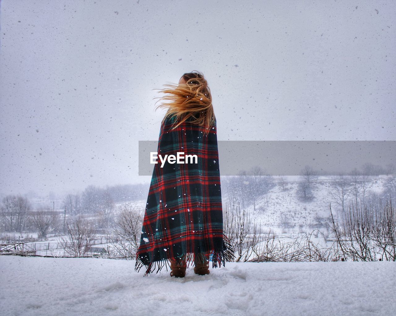 Woman wrapped in blanket standing on snow covered landscape against sky