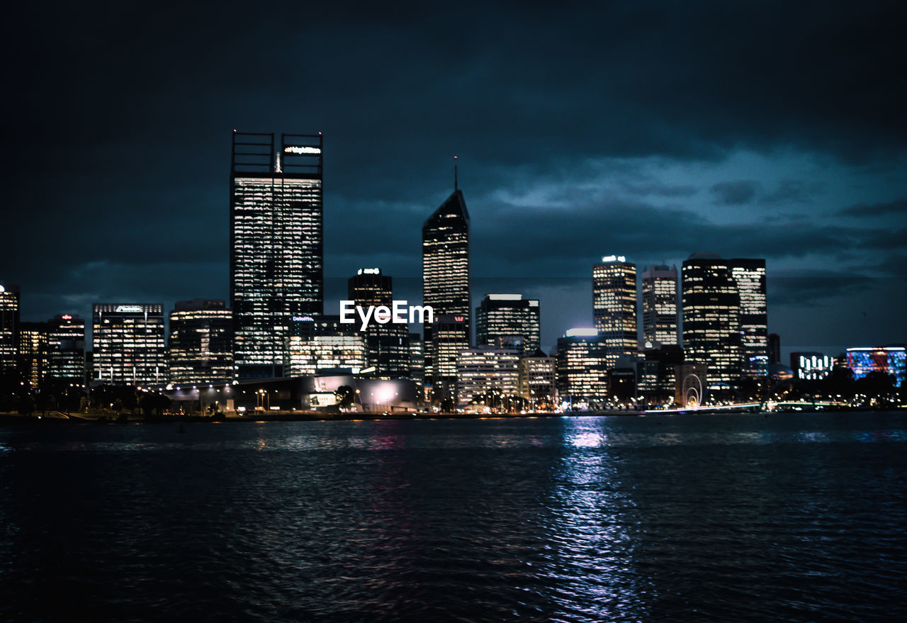 Illuminated skyscrapers by river against cloudy sky at night