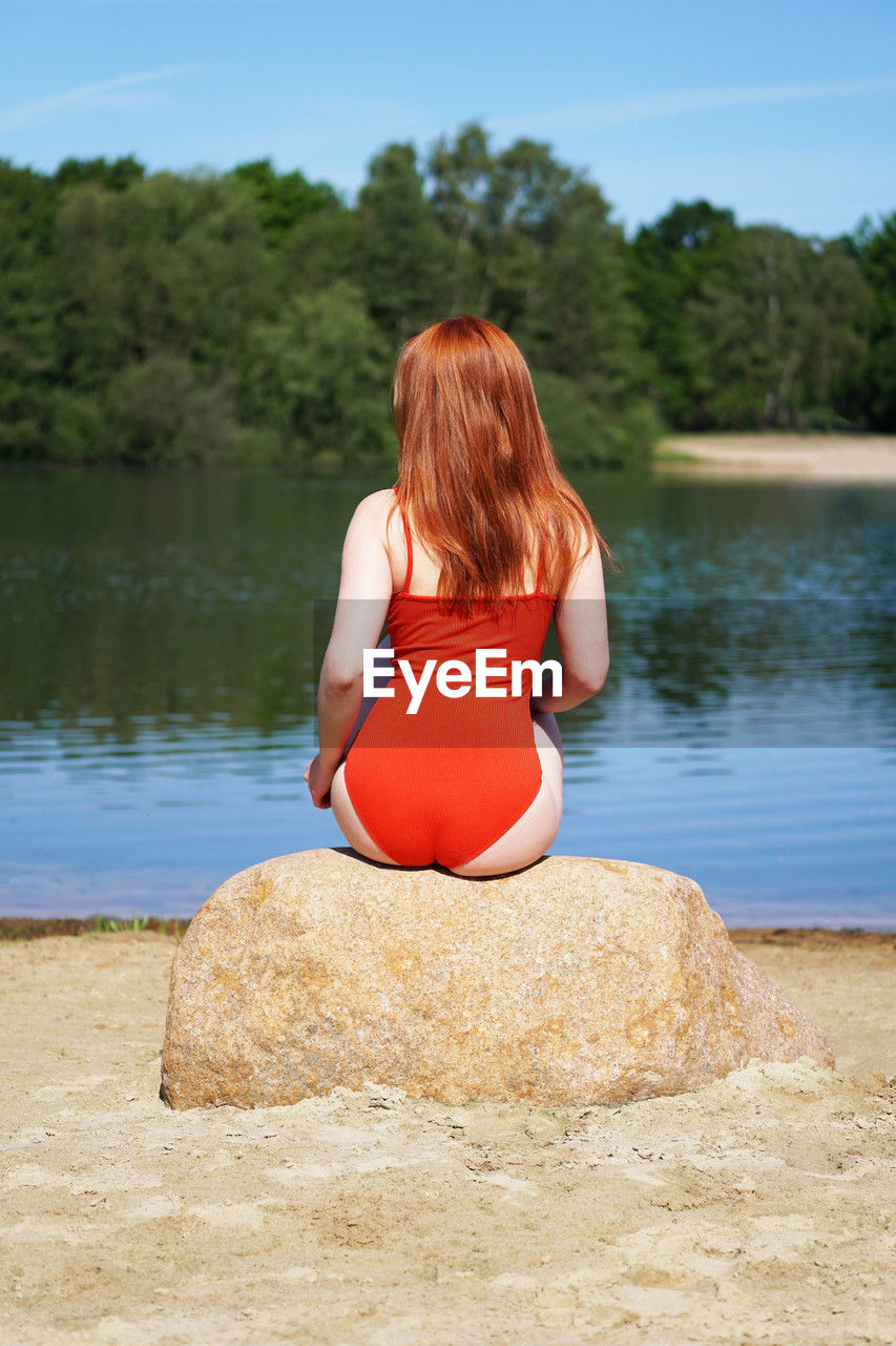 Rear view of young woman wearing red bathing suit sitting on rock on beach at idyllic lake