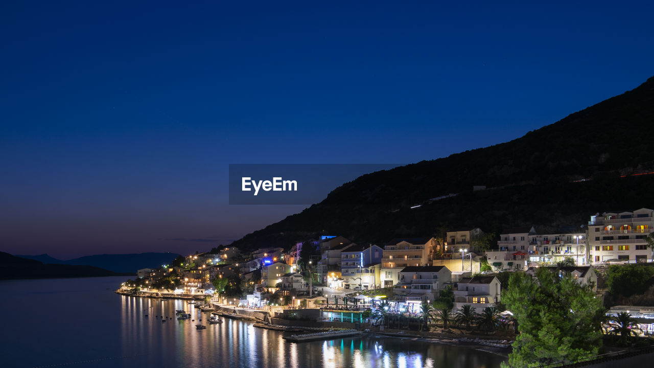 ILLUMINATED CITYSCAPE BY SEA AGAINST CLEAR BLUE SKY