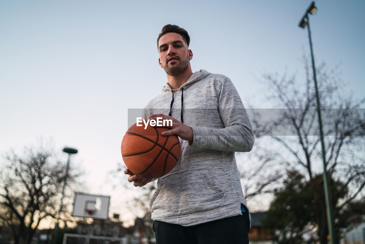 Portrait of young man holding basketball against sky